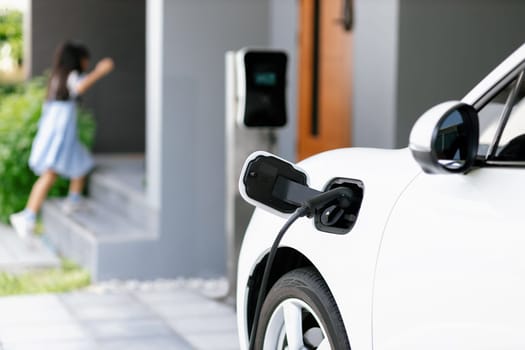 Focus EV car recharging battery at home charging station with blurred girl running in the background. Alternative clean energy driving vehicle for progressive lifestyle concept.