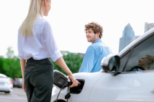 Progressive businessman and businesswoman with electric car connected to charging station before driving around city center. Eco friendly rechargeable car powered by alternative clean energy.