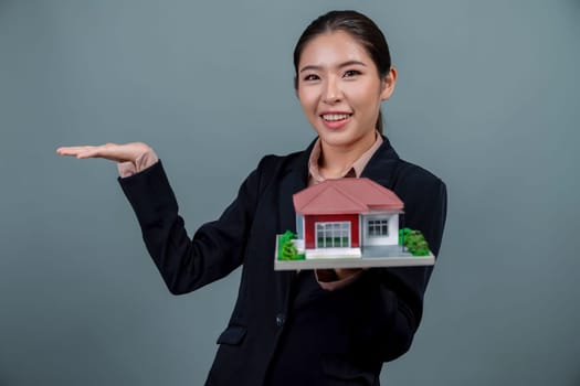 Young Asian woman wearing formal suit, holding and showcasing house model on isolated background. Ideal for housing advertisement, home insurance promotions, or customizable copy space. Enthusiastic