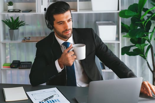 Manager of call center operator office sitting on his desk with his coffee while working on laptop. Office worker wearing headset and black suit working on customer support or telemarketing. fervent