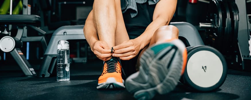 Sport male trainer tying his shoelace while sitting on the gym floor. The shot highlights his sporty shorts and emphasizes his dedication to fitness and healthy living