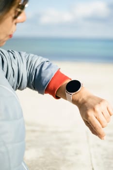 Cropped photo of a stylish woman checks notification on wearable digital wrist watch monitors the weather wears casual jacket poses on the street. Woman uses modern gadget on the street