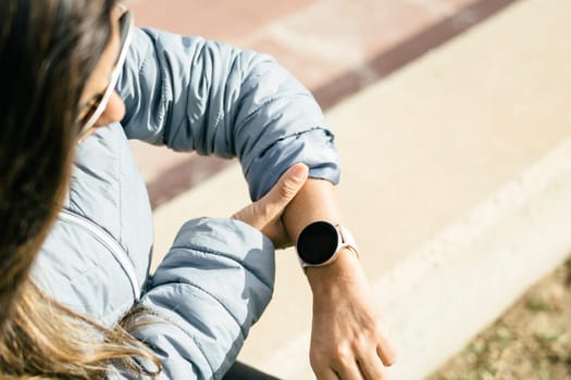 This stock photo shows a young Latina woman focused on her smart watch as she checks her heart rate. The image reflects the woman's commitment to her well-being and her interest in technology as a tool for monitoring her health. The blurred background highlights her figure, while the natural light accentuates her natural beauty. This image would be perfect for illustrating articles or posts related to personal care and technology in health.