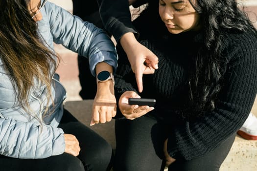 happy friends sharing multimedia content online with their smartphones and smartwatch, two beautiful Latina women sitting outdoors