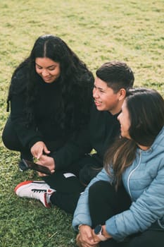 Hispanic family spending quality time together in a local park on a beautiful, sunny day. A teenage boy is sitting on the grass, holding his smartphone while looking away. His mother and sister are sitting next to him, looking at the camera and smiling.