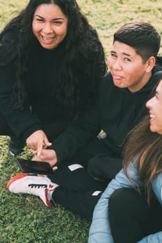 beautiful latin family laughing from watching smartphone videos, sitting on the green park two brothers and the mother on a sunny day.