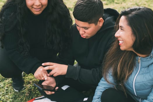 three people of hispanic-latino ethnicity sitting on the green grass of a park, with smart device. relaxed and comfortable as they entertain themselves with their screens, browsing social networks, dressed in casual clothes and their faces suggest satisfaction with their activity. it conveys a sense of modernity and connectivity that is common in today's digital age.