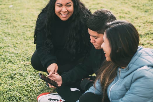 beautiful latin family laughing from watching smartphone videos, sitting on the green park two brothers and the mother on a sunny day.