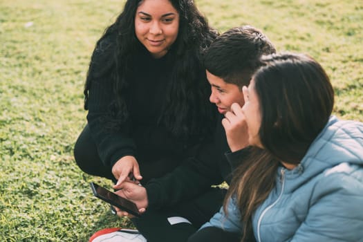 Hispanic family spending quality time together in a local park on a beautiful, sunny day. A teenage boy is sitting on the grass, holding his smartphone while looking away. His mother and sister are sitting next to him, looking at the camera and smiling