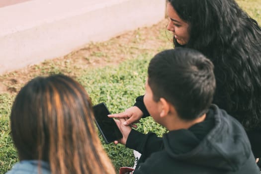 three Hispanic friends taking a break from their busy schedules and enjoying a sunny day in the park. The image is taken from behind and shows the group sitting on the grass, looking at a smartphone held by one of the young men. They seem to be engrossed in whatever is on the screen, with their heads close together.relaxing atmosphere. The photo emphasizes the importance of technology in modern social interactions, but also the need to take a break and enjoy some leisure time in the great outdoors.social media use, multiculturalism, and the balance between technology and nature.