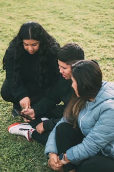 three people of hispanic-latino ethnicity sitting on the green grass of a park, with smart device. relaxed and comfortable as they entertain themselves with their screens, browsing social networks, dressed in casual clothes and their faces suggest satisfaction with their activity. it conveys a sense of modernity and connectivity that is common in today's digital age.