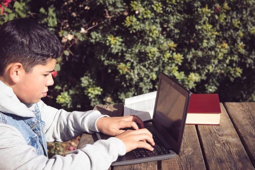 young man in denim jacket, sitting on wooden bench writing literature work on computer, books on table outside home in the park