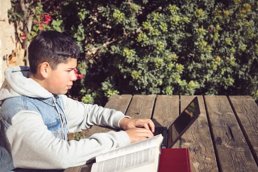 young latino student, working in the park with books and laptop on a sunny, cold day, sitting on a wooden bench