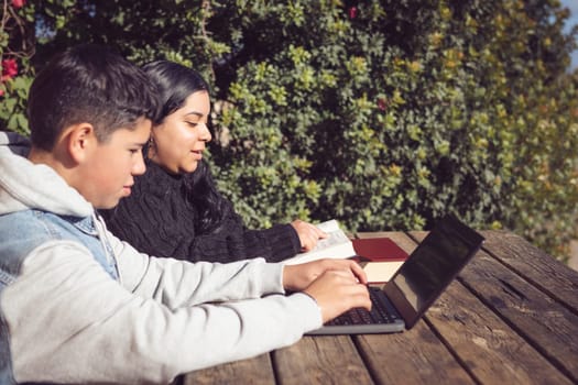 two young Latino students, outdoors, using laptops, sitting at a park table working with a computer