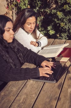 two beautiful young latinas working with books and laptop outdoors in city park sitting on a wooden bench on a sunny day