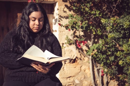 young latina girl sitting reading a book in the garden next to the country house
