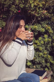 A Latina woman sits on a wooden bench in the garden, taking a sip of coffee while enjoying a moment of calm and relaxation. The natural beauty of the garden is appreciated in the background, creating a harmonious and peaceful atmosphere.