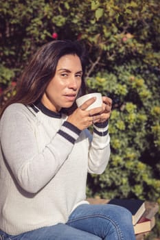 A Latina woman is sitting on a wooden bench in the garden, enjoying the fresh air while admiring the beauty of the flowers and plants. She holds a cup of coffee in her hand, while smiling at the natural surroundings