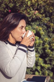 A Latina woman sits on a wooden bench in the middle of the garden, enjoying a moment of rest while taking a sip of coffee from her mug. Her face reflects a relaxed and content expression, while admiring the surrounding nature. The image conveys a sense of peace and tranquility, inviting the viewer to enjoy a moment of calm and contemplation. The natural environment of the garden, combined with the coffee mug, evokes a cozy and comforting atmosphere.