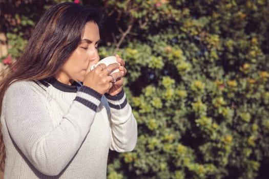 A Latina woman enjoys the garden from a wooden bench, taking a sip of coffee from her mug. The peace that the natural environment transmits is appreciated, in harmony with the pleasure of drinking a hot beverage.