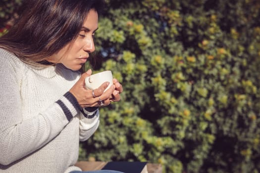 A Latina woman sits on a wooden bench in the garden, disconnecting from the outside world while taking a break to enjoy her coffee. The natural beauty of the garden conveys a sense of calm and peace, in harmony with the comforting sensation of drinking a hot beverage.