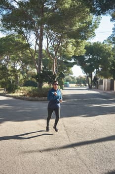 Mature fitness woman, between 30 and 50 years old, running, on the road, surrounded by pine trees, wearing sports glasses, bluetooth headphones, blue jacket, on a sunny winter day, Active Latin woman doing training, cardio workout, to burn calories