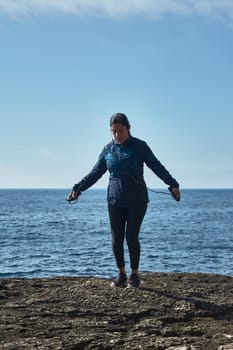 Middle-aged Latin woman jumping rope, warmed up for workout, burning calories and keeping fit, outdoors by the sea, blue sky, lovely sunny winter day, wearing bluetooth headphones,