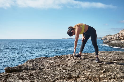 Middle-aged Latin woman, with yellow top, black leguin, doing stretching, gymnastics, warmed up for training, burning calories, keeping fit, outdoors by sea, blue sky, beautiful day, sunny winter, bluetooth headphones, smart watch, record training