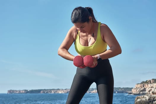 Middle-aged Latin woman, with yellow top, black leguin, with dumbbells, gymnastics, warmed up for training, burn calories, keep fit, outdoors by sea, blue sky, beautiful day, sunny winter, bluetooth headphones, smart watch, record the training