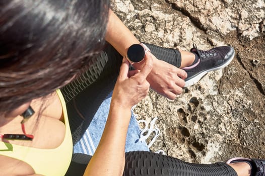 Latin woman, middle-aged, looking at app, smart watch, resting after gym session, wearing yellow top, black leggings, dumbbell, burning calories, staying fit, outdoors by the sea, blue sky, beautiful day, sunny winter, Bluetooth headphones,