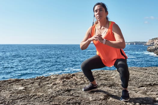 Latin woman, middle-aged, with orange top, black leggings, training, doing physical exercises, squats, burning calories, keeping fit, outdoors by the sea, blue sky, day, sunny winter, bluetooth headphones, smart watch, record the trainin