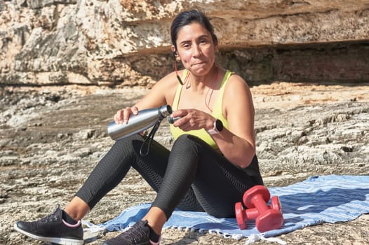 Latin woman, middle-aged, resting, regaining strength, eating, drinking water, after a gym session, burning calories, keeping fit, outdoors by the sea, wearing headphones and smart watch