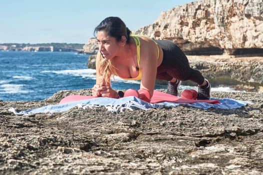 Latin woman, middle-aged, wearing sportswear, training, doing physical exercises, plank, sit-ups, climber's step, burning calories, keeping fit, outdoors by the sea, wearing headphones, smart watch