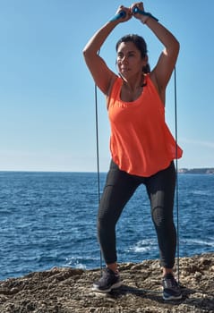 Latin woman, middle-aged, wearing sportswear, training, doing physical exercises, plank, sit-ups, climber's step, burning calories, keeping fit, outdoors by the sea, wearing headphones, smart watch