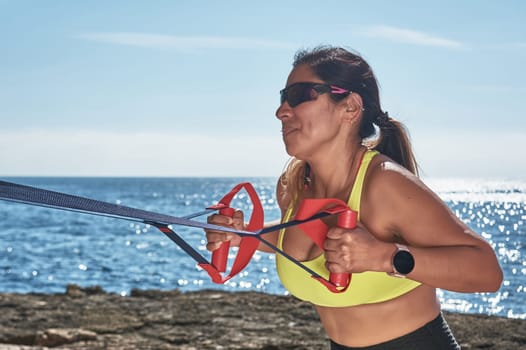 Latin woman, middle-aged, wearing sportswear, training, doing physical exercises, plank, sit-ups, climber's step, burning calories, keeping fit, outdoors by the sea, wearing headphones, smart watch