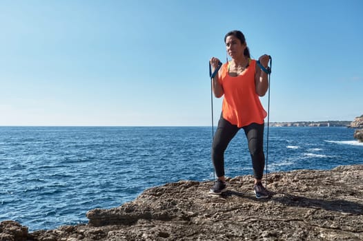 Latin woman, middle-aged, wearing sportswear, training, doing physical exercises, plank, sit-ups, climber's step, burning calories, keeping fit, outdoors by the sea, wearing headphones, smart watch