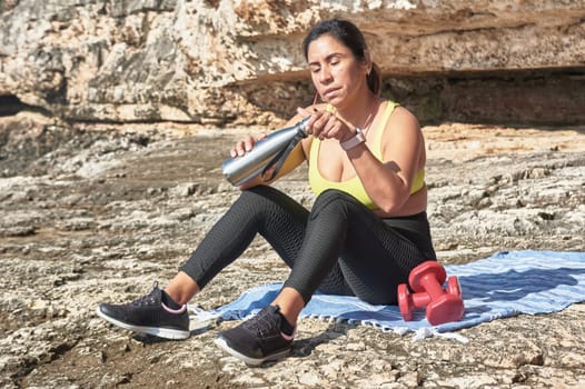 Latin woman, middle-aged, resting, regaining strength, eating, drinking water, after a gym session, burning calories, keeping fit, outdoors by the sea, wearing headphones and smart watch