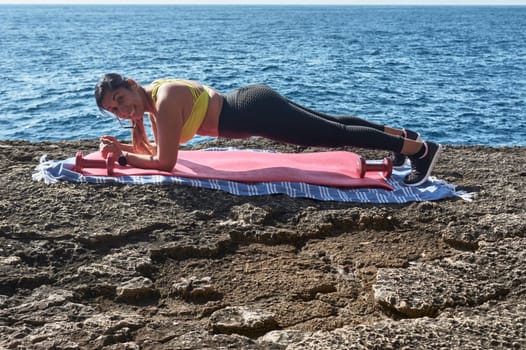 Latin woman, middle-aged, wearing sportswear, training, doing physical exercises, plank, sit-ups, climber's step, burning calories, keeping fit, outdoors by the sea, wearing headphones, smart watch