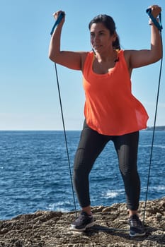 Latin woman, middle-aged, wearing sportswear, training, doing physical exercises, musculature, with elastic band, burning calories, keeping fit, outdoors by the sea, wearing headphones, smart watch