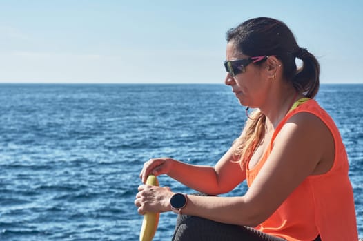 Latin woman, middle-aged, resting, regaining strength, eating, drinking water, after a gym session, burning calories, keeping fit, outdoors by the sea, wearing headphones and smart watch