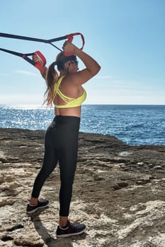 Latin woman, middle-aged, wearing sportswear, training, doing physical exercises, musculature, with elastic band, burning calories, keeping fit, outdoors by the sea, wearing headphones, smart watch