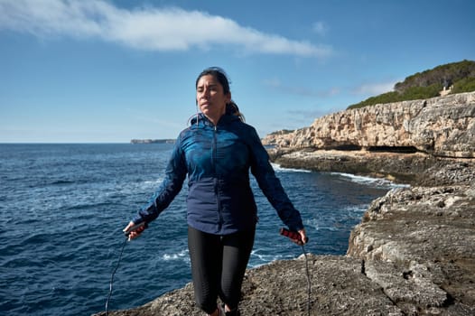 Latin woman, middle-aged, wearing sportswear, training, doing physical exercises, plank, sit-ups, climber's step, burning calories, keeping fit, outdoors by the sea, wearing headphones, smart watch