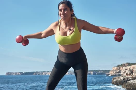 Latin woman, middle-aged, wearing sportswear, training, doing physical exercises, plank, sit-ups, climber's step, burning calories, keeping fit, outdoors by the sea, wearing headphones, smart watch