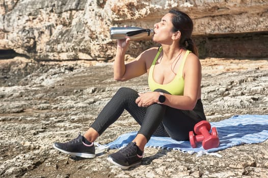 Latin woman, middle-aged, resting, regaining strength, eating, drinking water, after a gym session, burning calories, keeping fit, outdoors by the sea, wearing headphones and smart watch