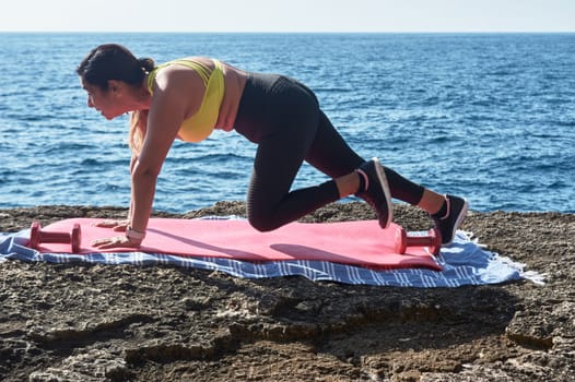 Latin woman, middle-aged, wearing sportswear, training, doing physical exercises, plank, sit-ups, climber's step, burning calories, keeping fit, outdoors by the sea, wearing headphones, smart watch