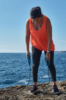 Latin woman, middle-aged, wearing sportswear, training, doing physical exercises, plank, sit-ups, climber's step, burning calories, keeping fit, outdoors by the sea, wearing headphones, smart watch