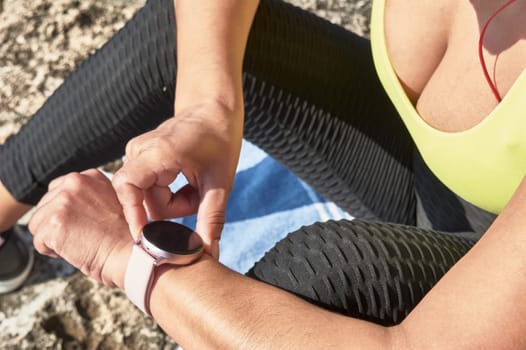 Latin woman, middle-aged, resting, regaining strength, eating, drinking water, after a gym session, burning calories, keeping fit, outdoors by the sea, wearing headphones and smart watch