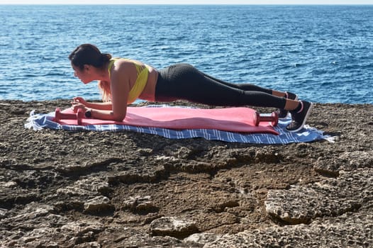 Latin woman, middle-aged, wearing sportswear, training, doing physical exercises, plank, sit-ups, climber's step, burning calories, keeping fit, outdoors by the sea, wearing headphones, smart watch