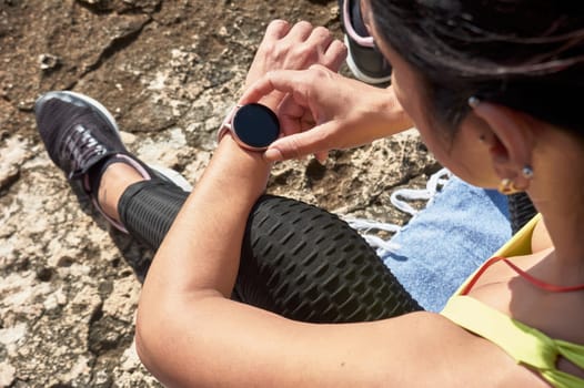 Latin woman, middle-aged, resting, regaining strength, eating, drinking water, after a gym session, burning calories, keeping fit, outdoors by the sea, wearing headphones and smart watch