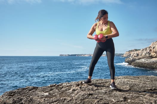 FITNESS WOMAN IN SPORTS SET TRAINING WITH ELASTIC BAND, WEIGHTS, GYM EXERCISES, IN FRONT OF THE WATER.
